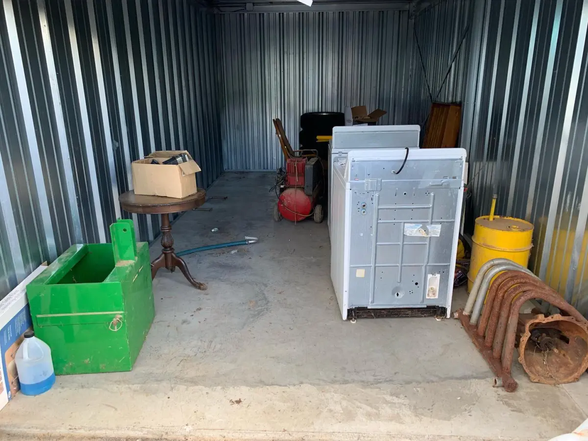 A storage unit with various items, including a green toolbox, a wooden table with a cardboard box, old washing machines, a yellow barrel, and rusted metal pipes.
