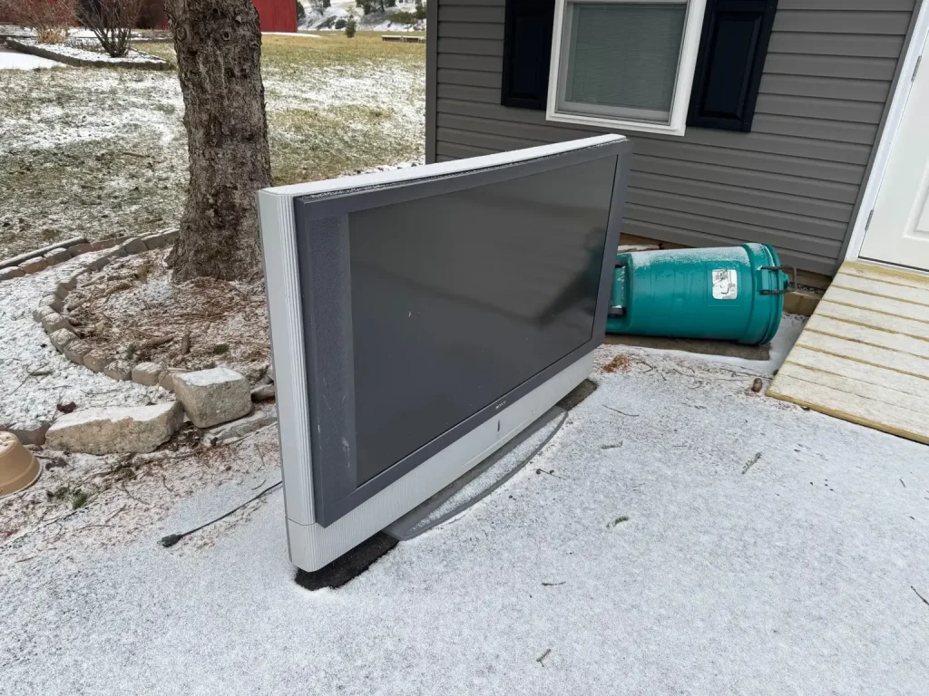 A large flat-screen TV lying on its back outdoors on a frosty surface, with a propane tank and a house entrance in the background.