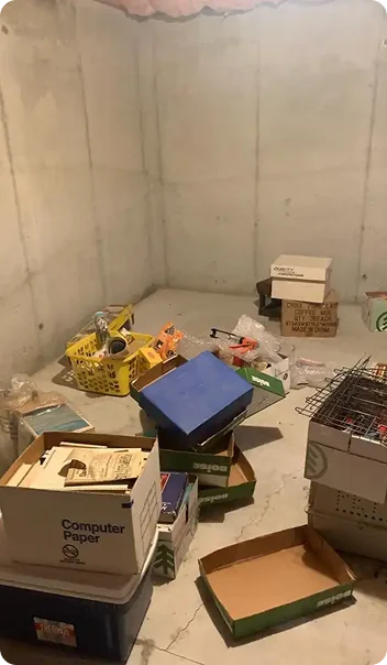 A cluttered basement storage area with stacked boxes, shelves, and various tools and items scattered on the floor.
