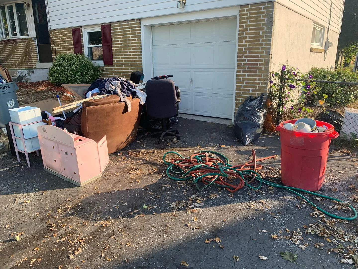 A cluttered driveway in front of a garage with assorted furniture, boxes, a chair, and tangled garden hoses scattered on the ground. A red trash bin and a black garbage bag sit nearby.